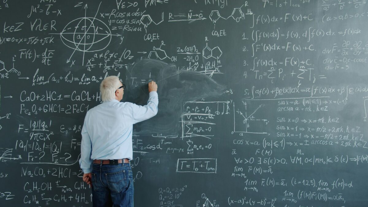Mature man physicist writing formulas on blackboard with chalk indoors at school busy with solving scientific problem.