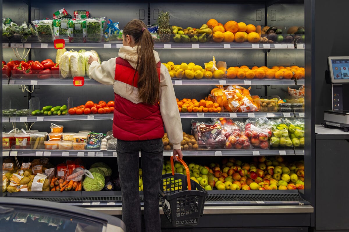 Woman Choosing Fresh Produce in Grocery Store. Female shopper with basket selecting vegetables in a grocery store produce aisle filled with colorful fruits, fresh food, and healthy choices